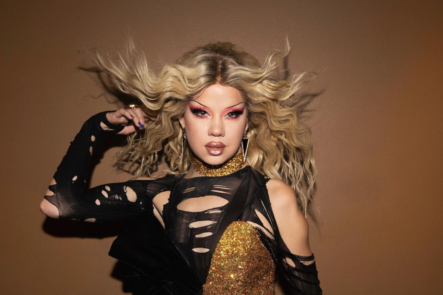 Willow Pill poses in a dramatic black and gold drag outfit, with voluminous blond hair and bold makeup.
