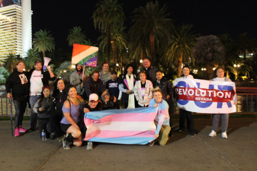 A group holds trans and queer flags during Las Vegas TransPride 2022 on the Strip, standing near The Mirage at night.