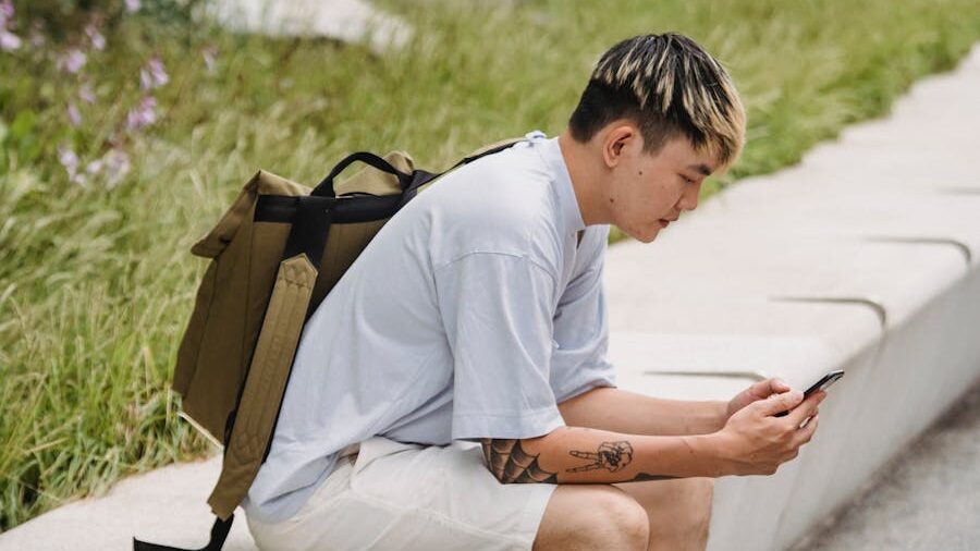 Young person with short hair and backpack sitting on a stone bench, looking at their phone in an outdoor park setting.