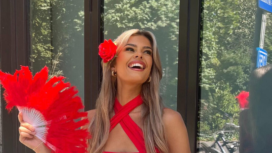 Smiling Rikkie Kollé wearing a red outfit and flower, seated by a table at Antwerp Pride 2024.