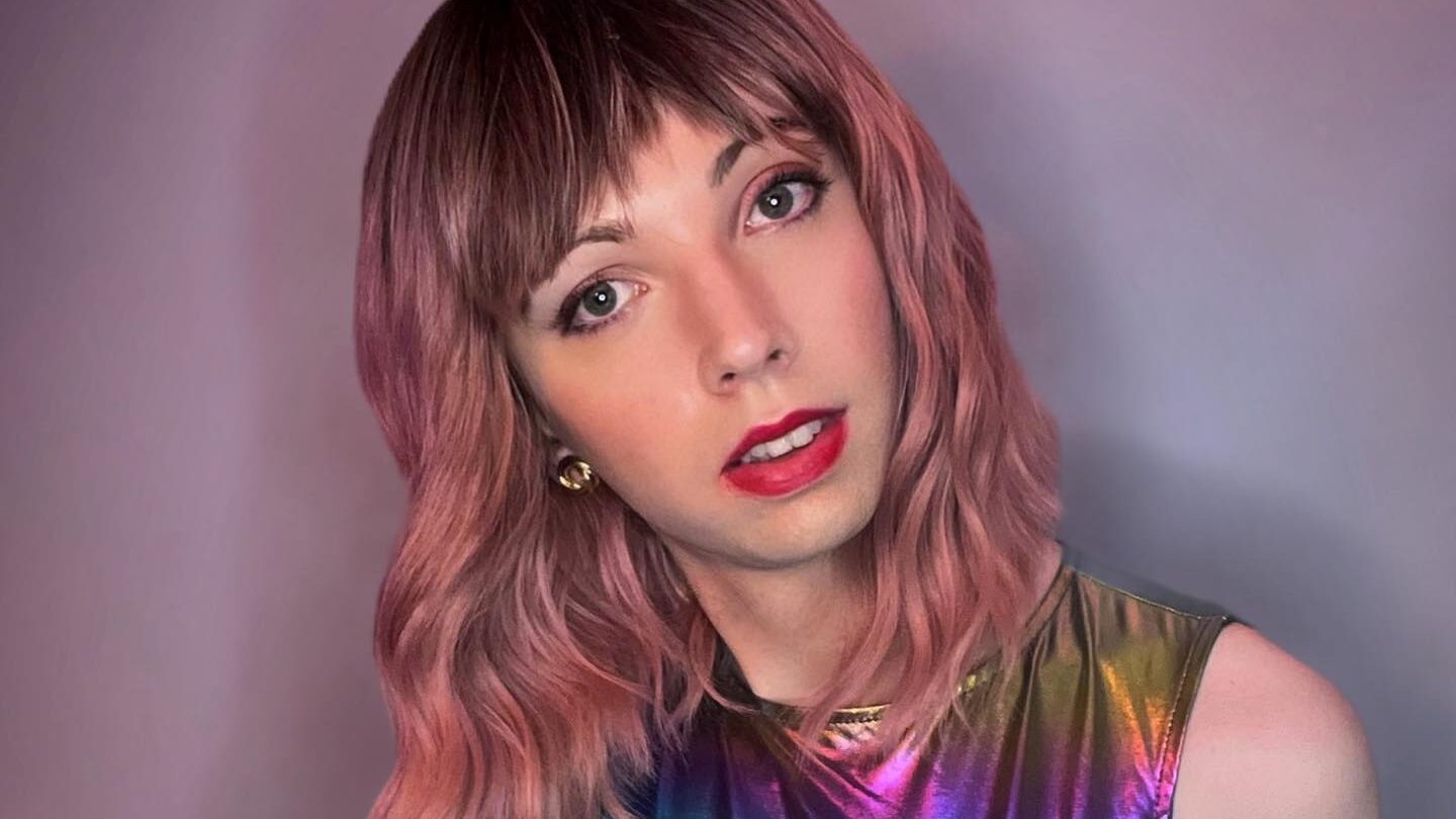 Rosie, wearing a shiny rainbow-colored iridescent dress, with shoulder-length pink wavy hair, striking a confident pose against a neutral background.