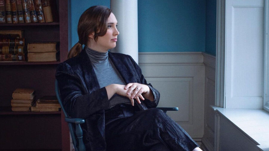 Sarah McBride assise près d’une fenêtre de l’Old State House à Dover, Delaware, vêtue d’un costume en velours sombre et regardant dehors avec réflexion.