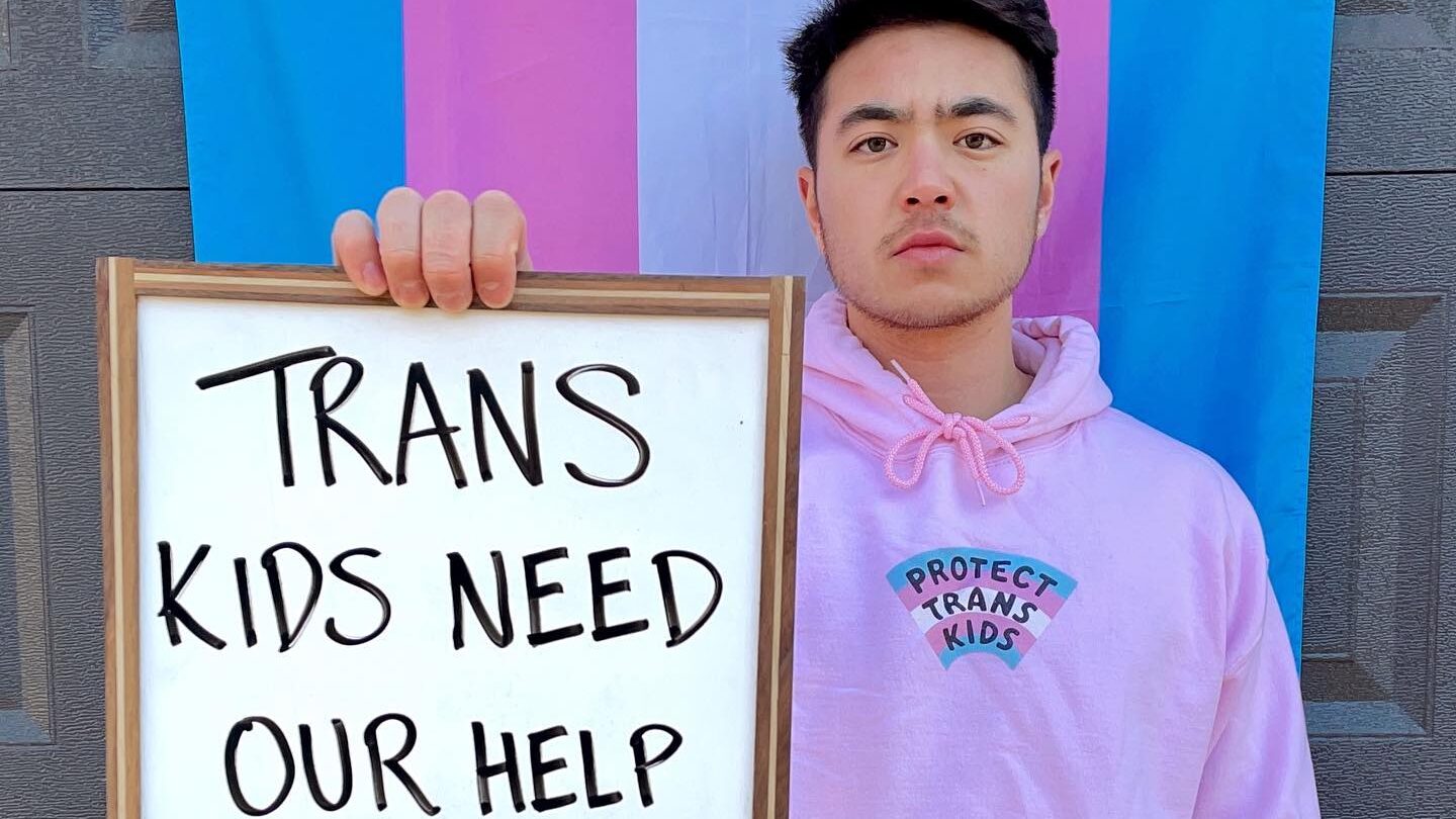 Schuyler Bailar stands in front of a trans flag, holding a sign that reads “Trans kids need our help now!!”.