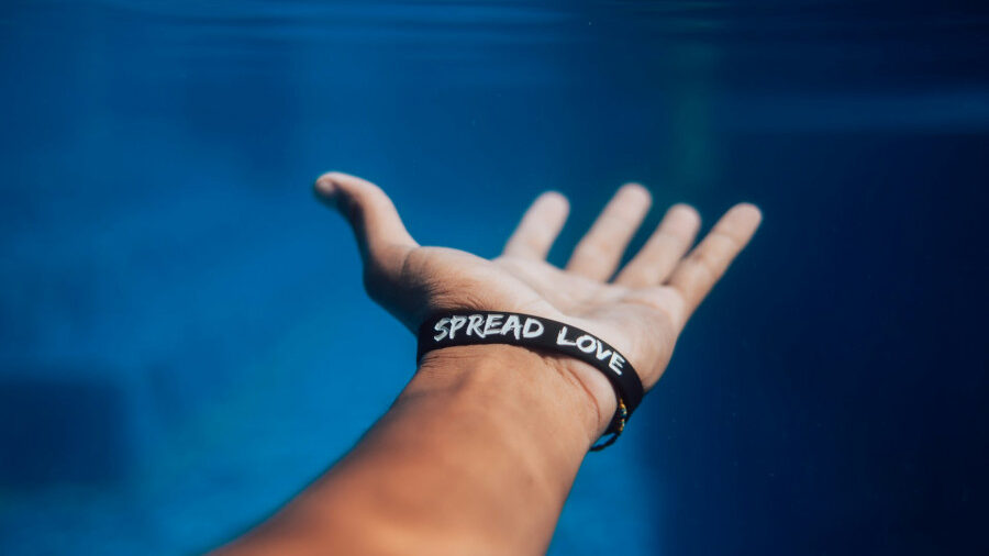 Close-up of a hand underwater, palm open, wearing a black bracelet with the white message “SPREAD LOVE.”