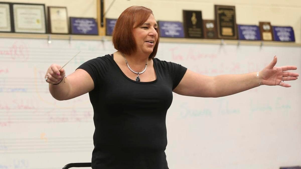Stephanie Byers stands in front of a whiteboard, arms outstretched, conducting a school music ensemble in a classroom.