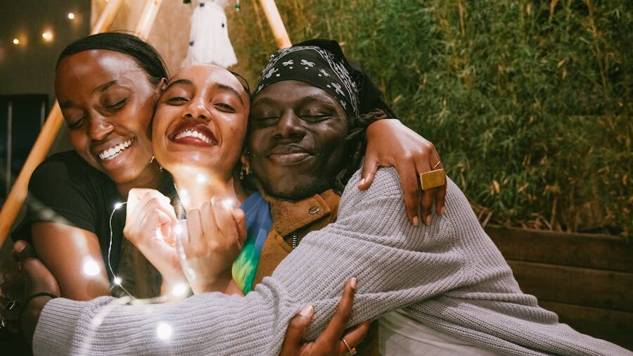 Three smiling people in a tight hug, surrounded by warm fairy lights, sharing a joyful and intimate moment.