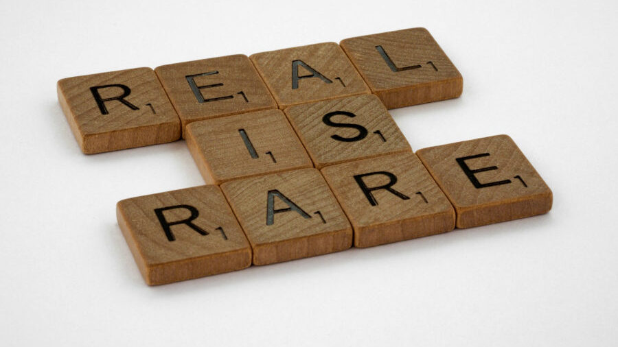 Scrabble-style wooden tiles arranged on a white surface spelling the phrase “REAL IS RARE” in block letters.