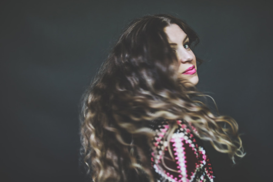 A trans woman with long wavy hair wearing a patterned outfit turns toward the camera with a confident expression, her hair flowing in motion.