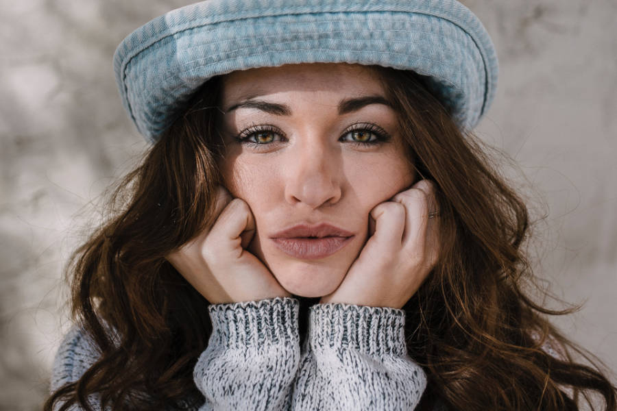 Trans woman from Boston in a blue hat and sweater, gazing into the camera with her chin resting on her hands.