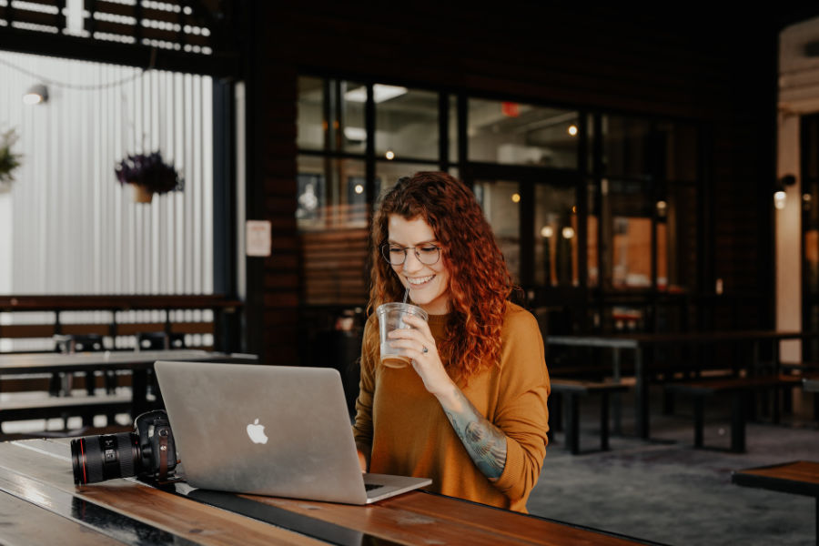 A trans woman with curly hair and glasses works on a laptop and smiles while drinking at a café with a camera nearby.
