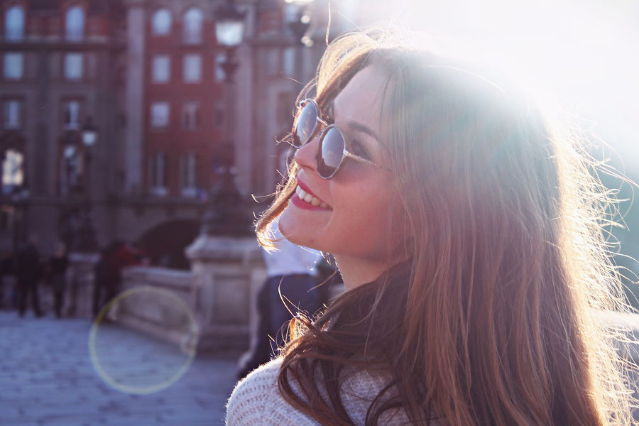 Trans woman smiling in the sun wearing sunglasses, with long hair backlit by sunlight in Fresno.