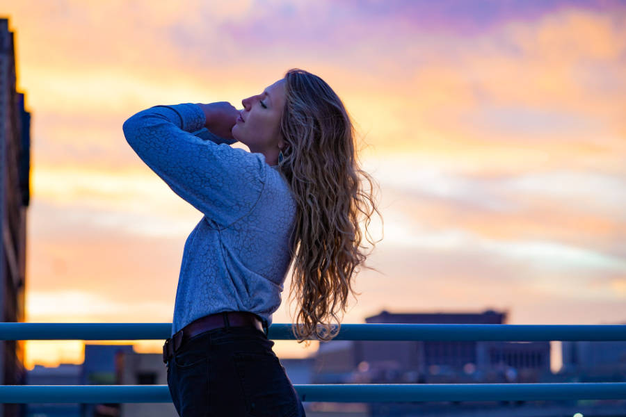 Transgender woman standing on a rooftop in Milwaukee during sunset, facing upward with long hair and casual outfit.