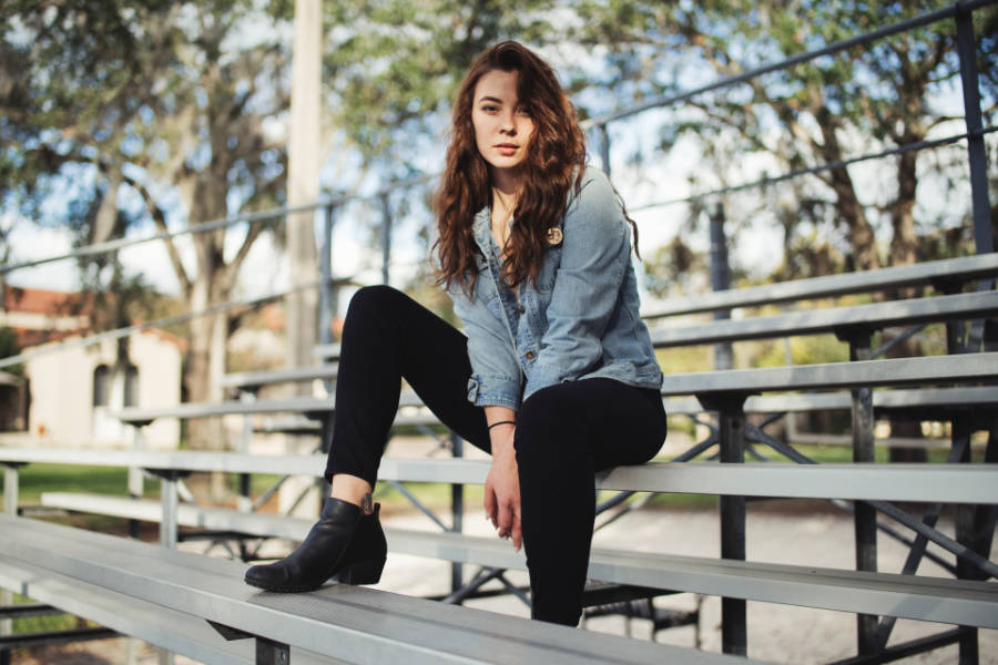 Trans woman from Orlando sitting confidently on bleachers, wearing a denim jacket, black pants, and boots.