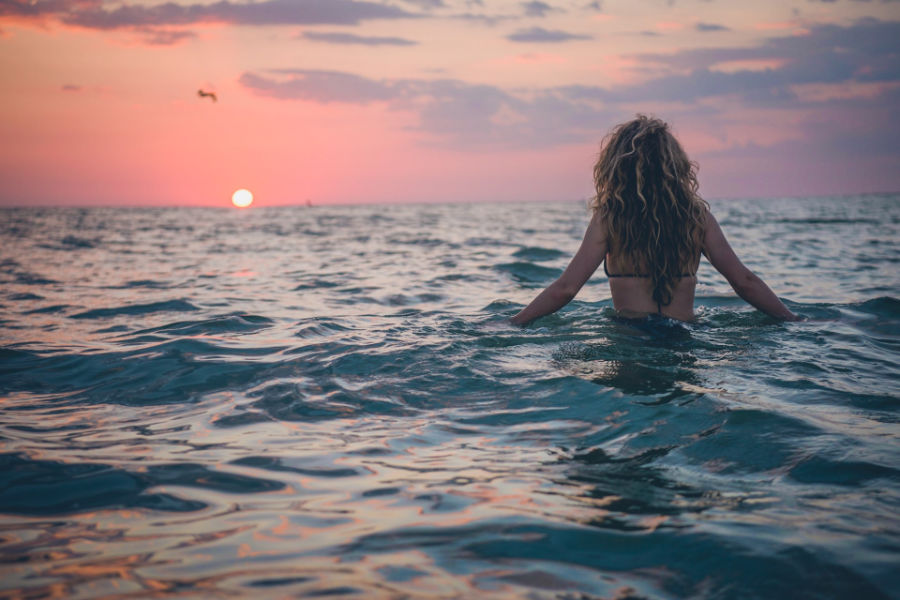 Trans woman with curly hair standing waist-deep in the sea, watching the sun set over the calm horizon.