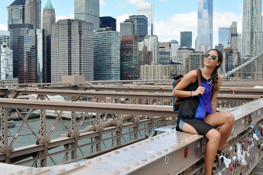 A trans woman sits on a bridge railing in NYC, wearing sunglasses and a braid, with skyscrapers in the background.