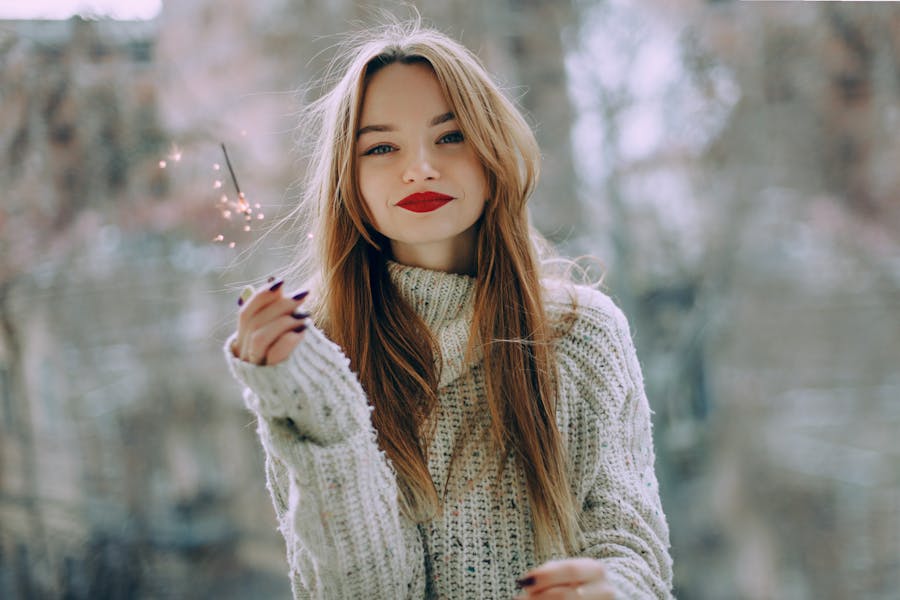 Smiling trans woman wearing a thick white sweater and red lipstick, holding a small sparkler in cold weather.