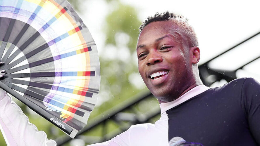 Todrick Hall smiling and holding a rainbow Progress Pride fan at an outdoor Pride event, showing support for Black trans lives.