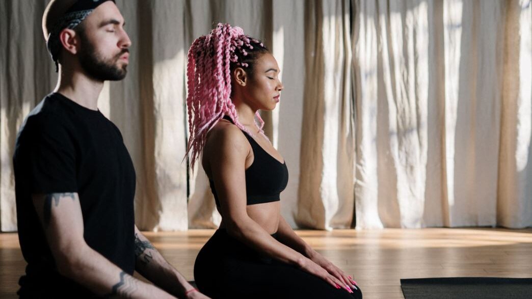 Mujer con trenzas rosadas y hombre con una banda en la cabeza meditando juntos.