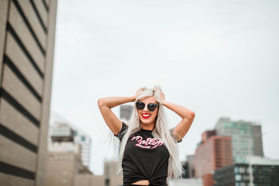 Confident trans woman in black t-shirt poses on a city rooftop, hands in hair, with tall buildings behind her.