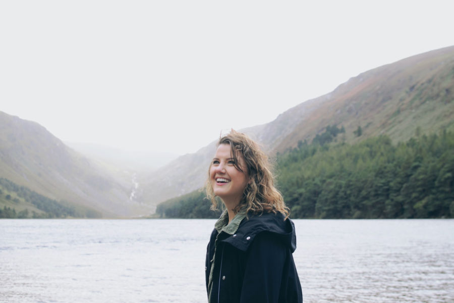 A smiling trans woman stands near a peaceful lake in Ireland, surrounded by mountains and evergreen forests.