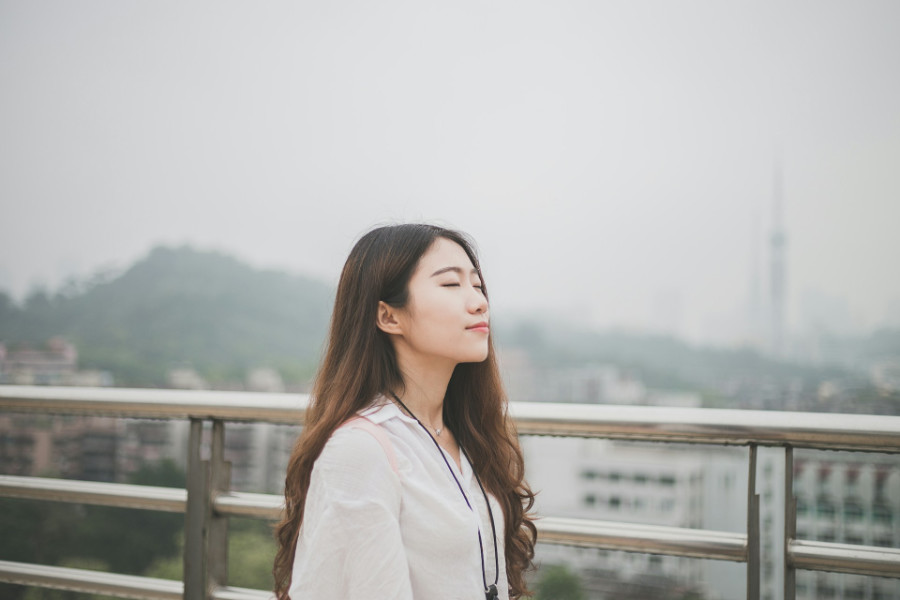 Side profile of a trans woman with long hair and eyes closed, standing on a rooftop with city view.