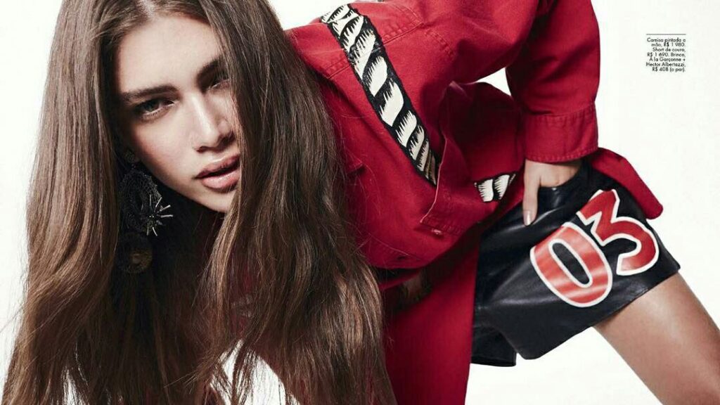 Valentina Sampaio wearing a red top and black shorts, leaning forward during a fashion shoot.