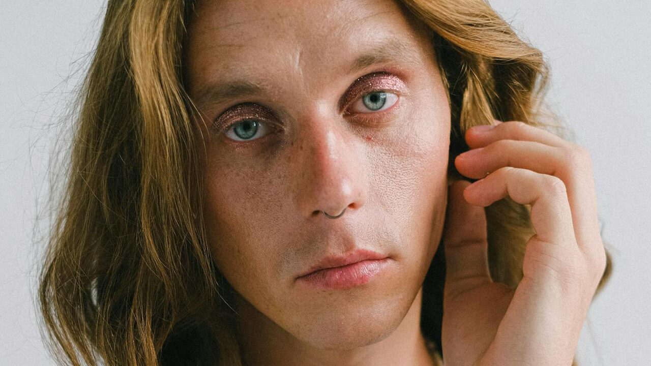 Una persona de ojos azules, con pecas y maquillaje natural se toca suavemente el rostro.