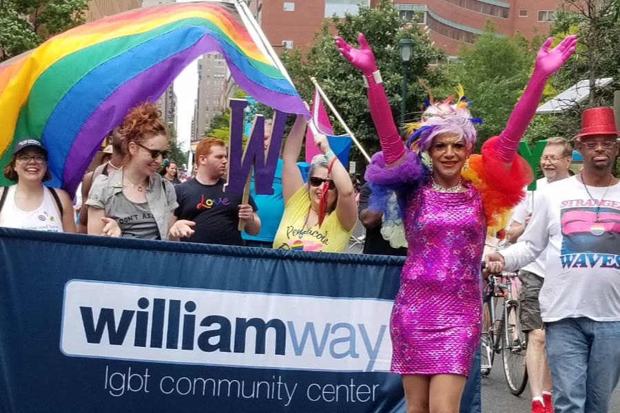 Parade group with a William Way banner, colorful costumes, and rainbow umbrellas walking through city streets.