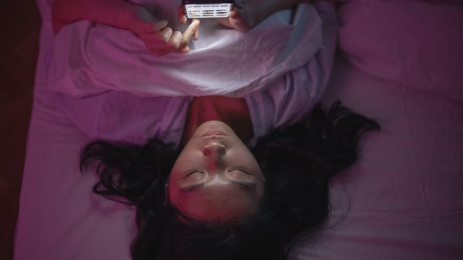 Woman lying on her back in bed in the dark, holding a phone that lights her face as she reads messages.