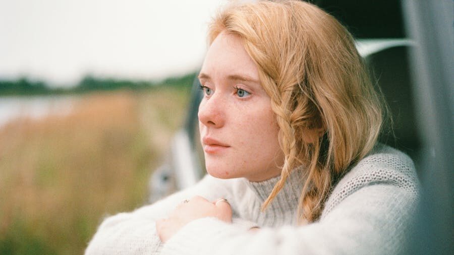 Blonde woman in a white sweater leaning on a car window, gazing thoughtfully toward a blurred natural landscape.