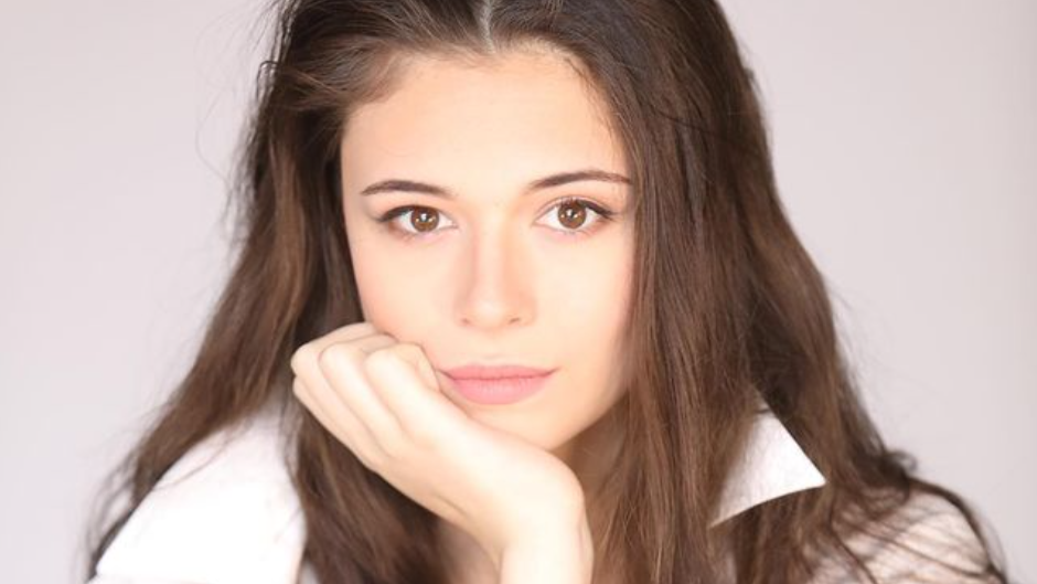 Nicole Maines poses in a white shirt with long brown hair, hand resting on her chin, against a soft neutral background.